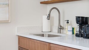Eine Küche mit weißer Arbeitsplatte, Spüle aus Edelstahl, Holzschränken, einer Kaffeemaschine und Gläsern auf Holzregalen.