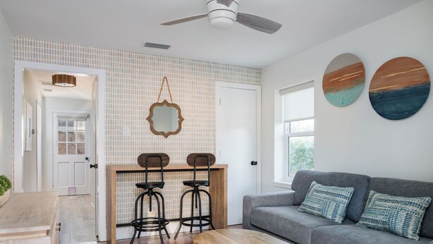 Ein modernes Wohnzimmer mit einem grauen Sofa, zwei schwarzen Barhocker, einem Holztisch und einem Deckenventilator.