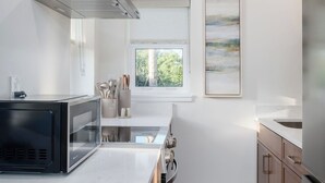 Eine moderne Küche mit Mikrowelle, Herd und einer Edelstahl-Dunstabzugshaube. Ein Fenster bietet Blick auf Grünflächen, und an der Wand hängt ein gerahmtes abstraktes Gemälde.
