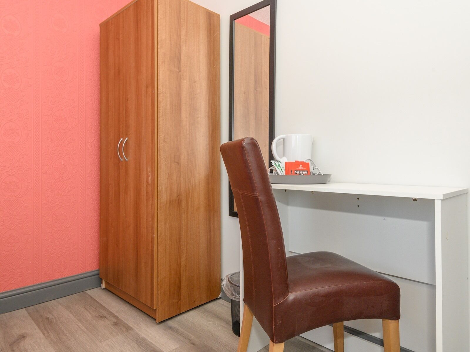 A room with a wooden wardrobe, a white desk, a brown chair, and a trash bin.