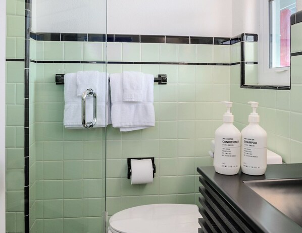 A bathroom with green tiles, a glass shower enclosure, a white toilet, and two bottles of shampoo and conditioner on a counter.