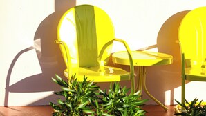 Two yellow chairs with a small table between them, positioned on a terrace with a white wall in the background and some green plants in the foreground. - The Castillo Queen Studio -5 min walk to beach! (St. Augustine)