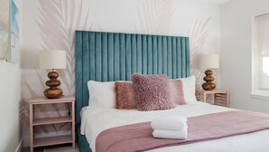 Een netjes ingerichte slaapkamer met een blauwgroen gestoffeerd hoofdeinde, twee nachtkastjes met lampen, en een bed met een roze plaid.