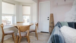 Een moderne woonkamer met een ronde houten tafel en stoelen, een donkerblauwe bank met een zachte witte plaid, en een groot raam met zonwering.
