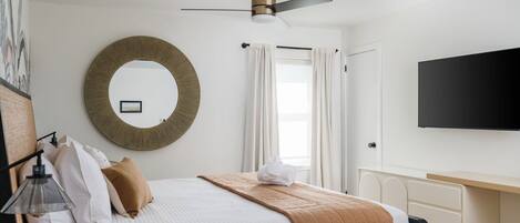 A hotel room with a bed, a round mirror, a ceiling fan, a flat-screen TV, and a desk with drawers.
