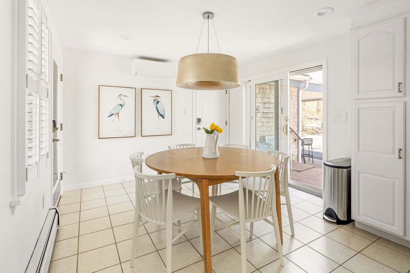 A dining area with a round wooden table, white chairs, a hanging light fixture, and two framed bird paintings on the wall.