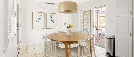 A dining area with a round wooden table, white chairs, a hanging light fixture, and two framed bird paintings on the wall.
