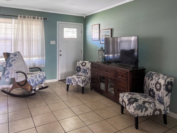 Un salon comprenant un fauteuil à bascule, deux fauteuils assortis, un téléviseur posé sur un meuble en bois et une porte avec une fenêtre.