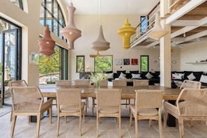 Un espace repas avec une grande table et des chaises en osier, des luminaires suspendus et une vue sur l’extérieur.