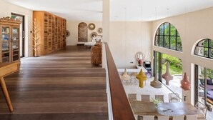 A spacious interior with a wooden floor, a dining area with a table and chairs, and large windows offering a view of the outdoors.