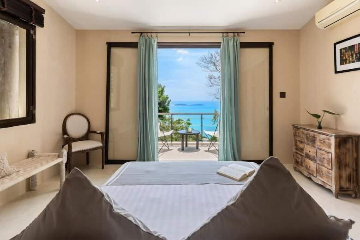 A bedroom with a bed, a chair, a small table, a cabinet, and a view of the sea.