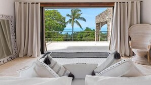 A room with a view of a palm tree and ocean, a balcony with a stone structure, and a bed with white bedding and striped pillows.