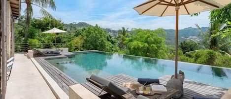 An infinity pool with lounge chairs, a table, and an umbrella, surrounded by lush greenery and hills.