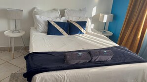 Una cama bien tendida con sábanas blancas y dos almohadones decorativos, un plaid azul oscuro y dos toallas con texto bordado. Hay dos mesitas de noche con lámparas, una mesita redonda pequeña y una pared de acento azul.