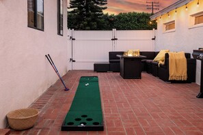 Área de pátio com mesa de shuffleboard verde, móveis de exterior e cerca branca.