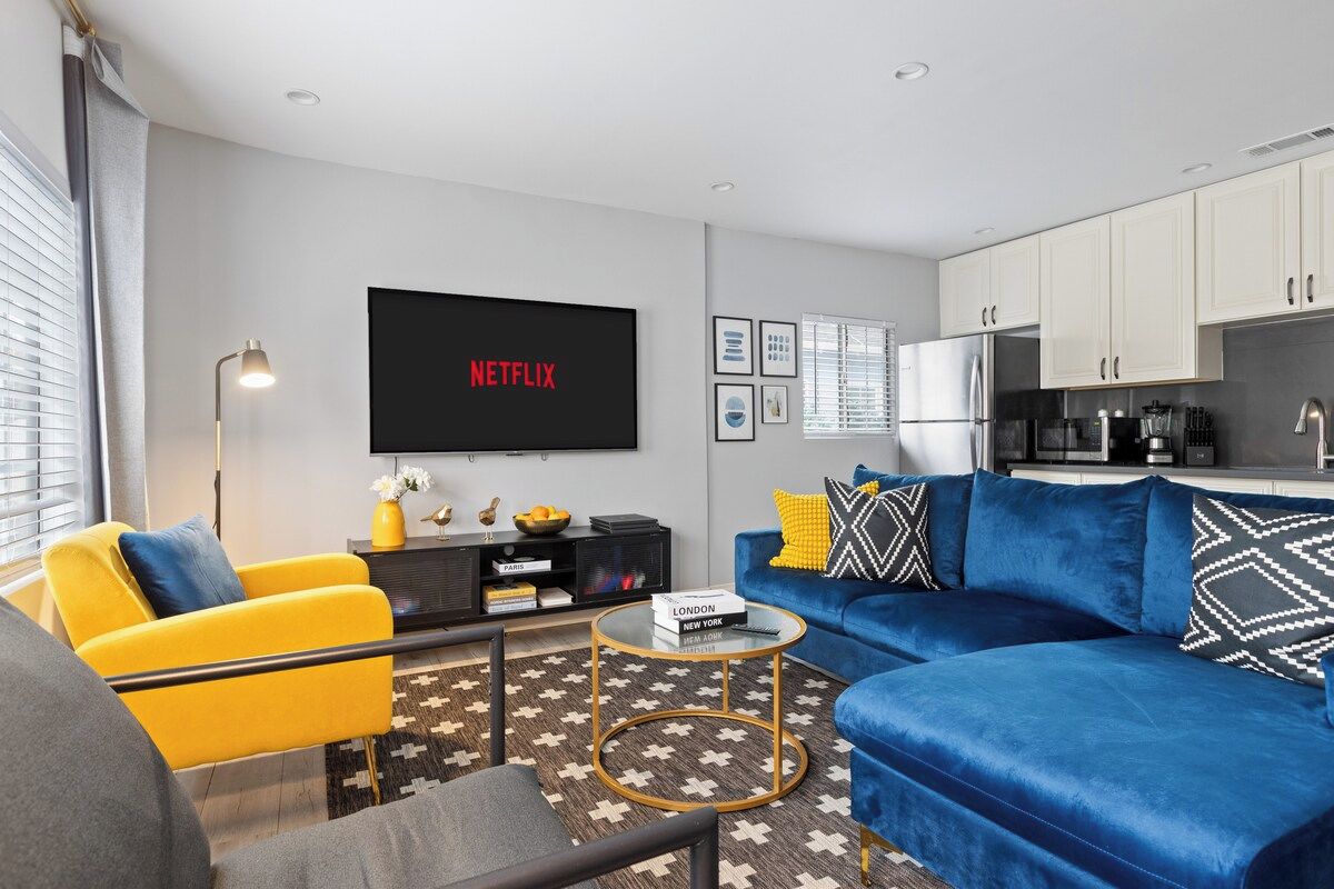 Een moderne woonkamer met een blauwe bank, een gele fauteuil, een salontafel, een flatscreen-tv en een keuken met kasten en apparatuur.