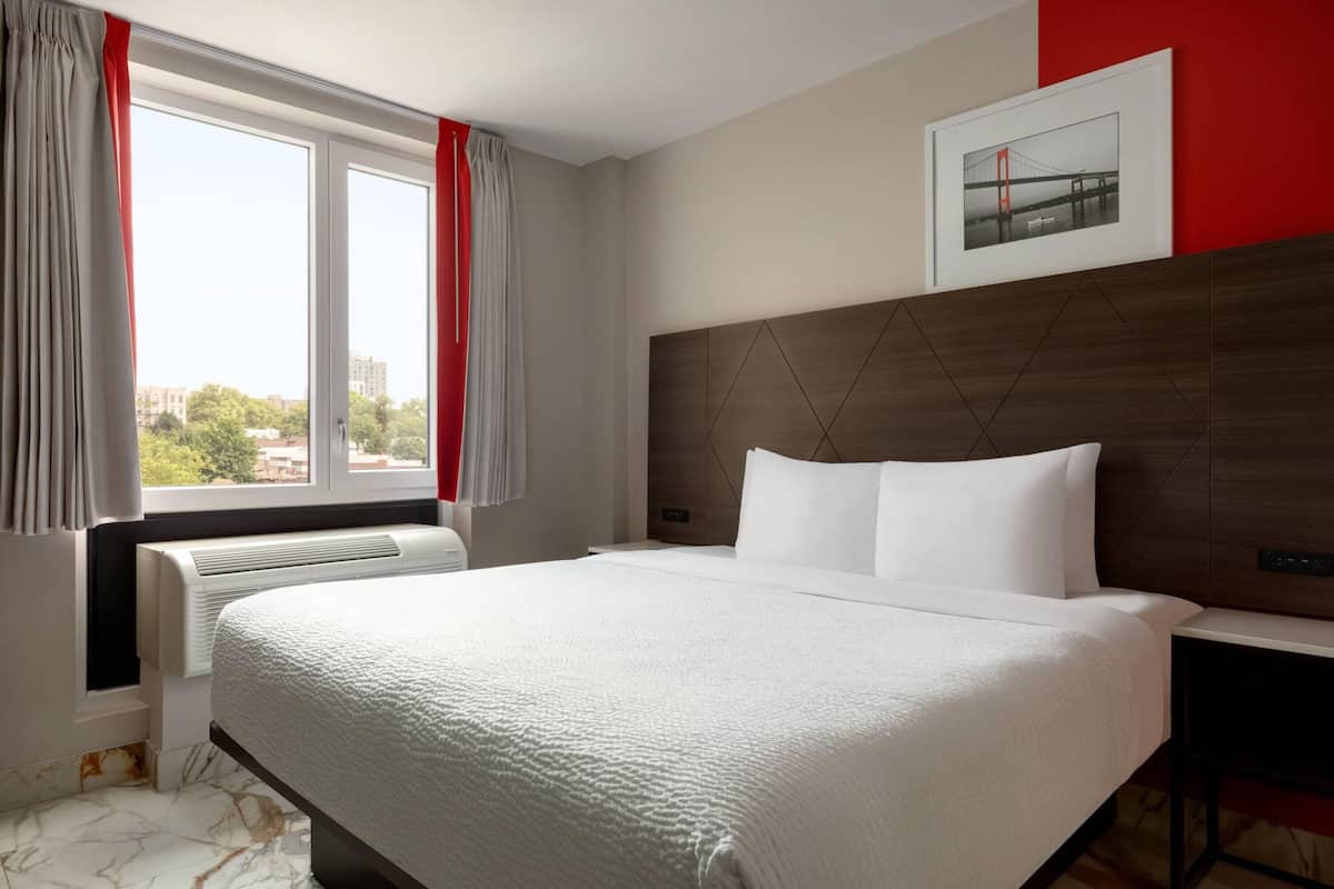 Ein Hotelzimmer mit einem großen Bett, einem Nachttisch, einem gerahmten Bild an der Wand und einem Fenster mit Vorhängen.