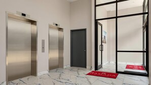 Un couloir avec deux ascenseurs, une porte et un tapis rouge sur lequel est inscrit le mot « RANADA ».