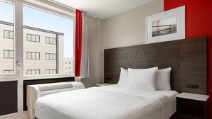 Une chambre d’hôtel avec un grand lit, une tête de lit ornée de motifs géométriques, une table de chevet et une fenêtre donnant sur des immeubles.