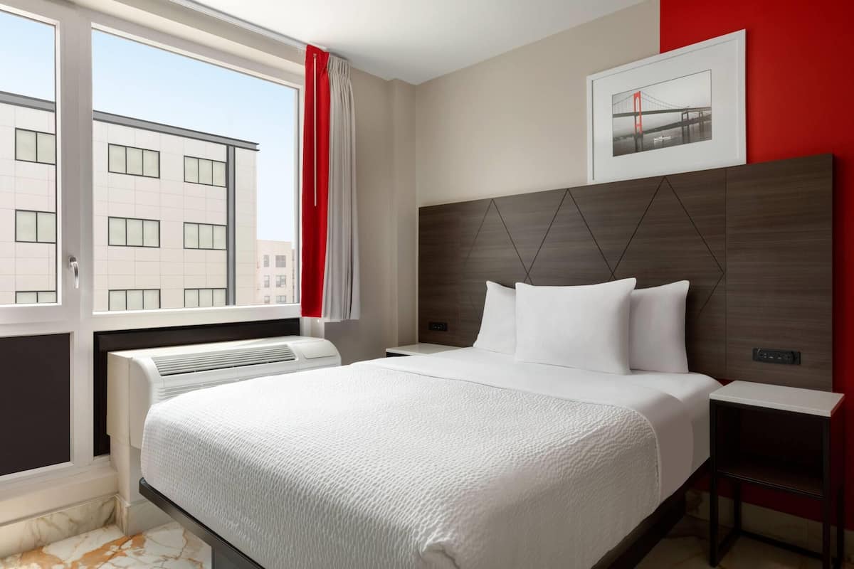 Ein Hotelzimmer mit einem großen Bett, einem Kopfende mit geometrischen Mustern, einem Nachttisch und einem Fenster mit Blick auf Gebäude.