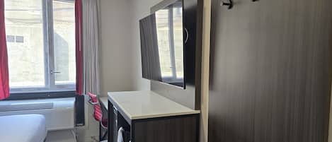 A hotel room with a bed, a desk with a mirror, a window with curtains, and a door.