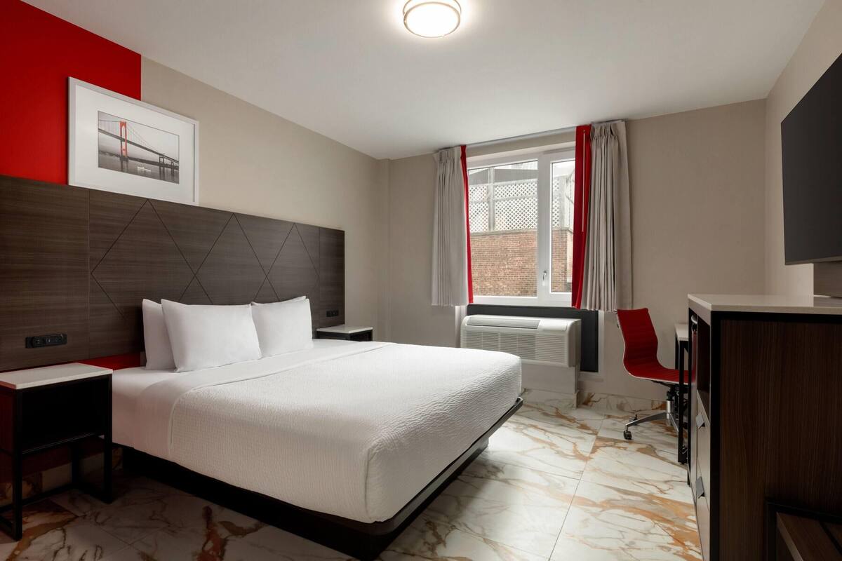 Ein Hotelzimmer mit einem großen Bett, einem Nachttisch, einem Schreibtisch, einem Stuhl, einem Fernseher und einem Fenster mit Vorhängen.