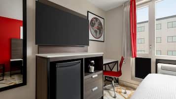 A hotel room with a bed, a desk, a chair, a small refrigerator, a large mirror, and a window with curtains.