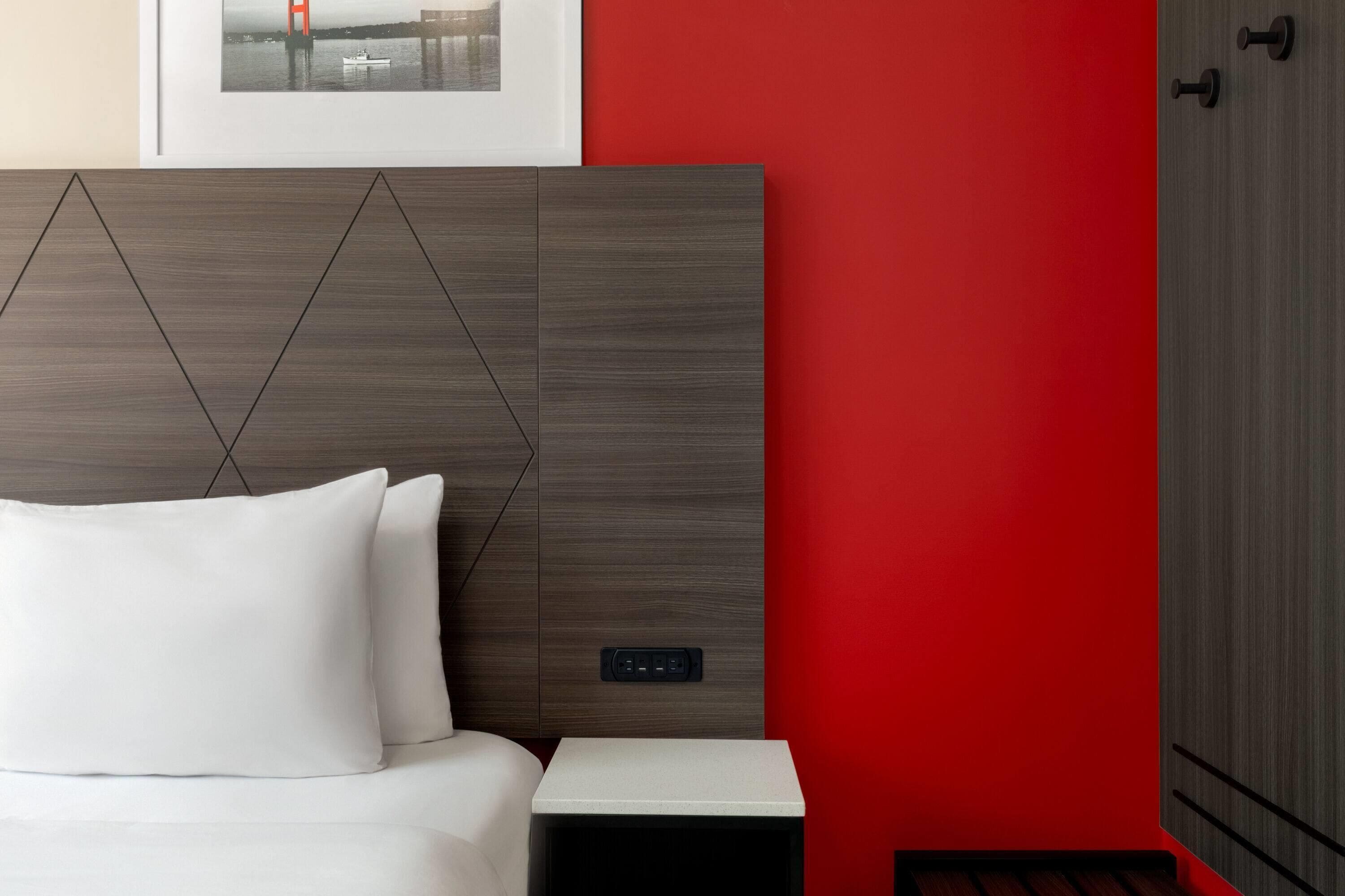 Habitación de hotel con cama, mesita de noche, toma de corriente y una pared de acento roja.