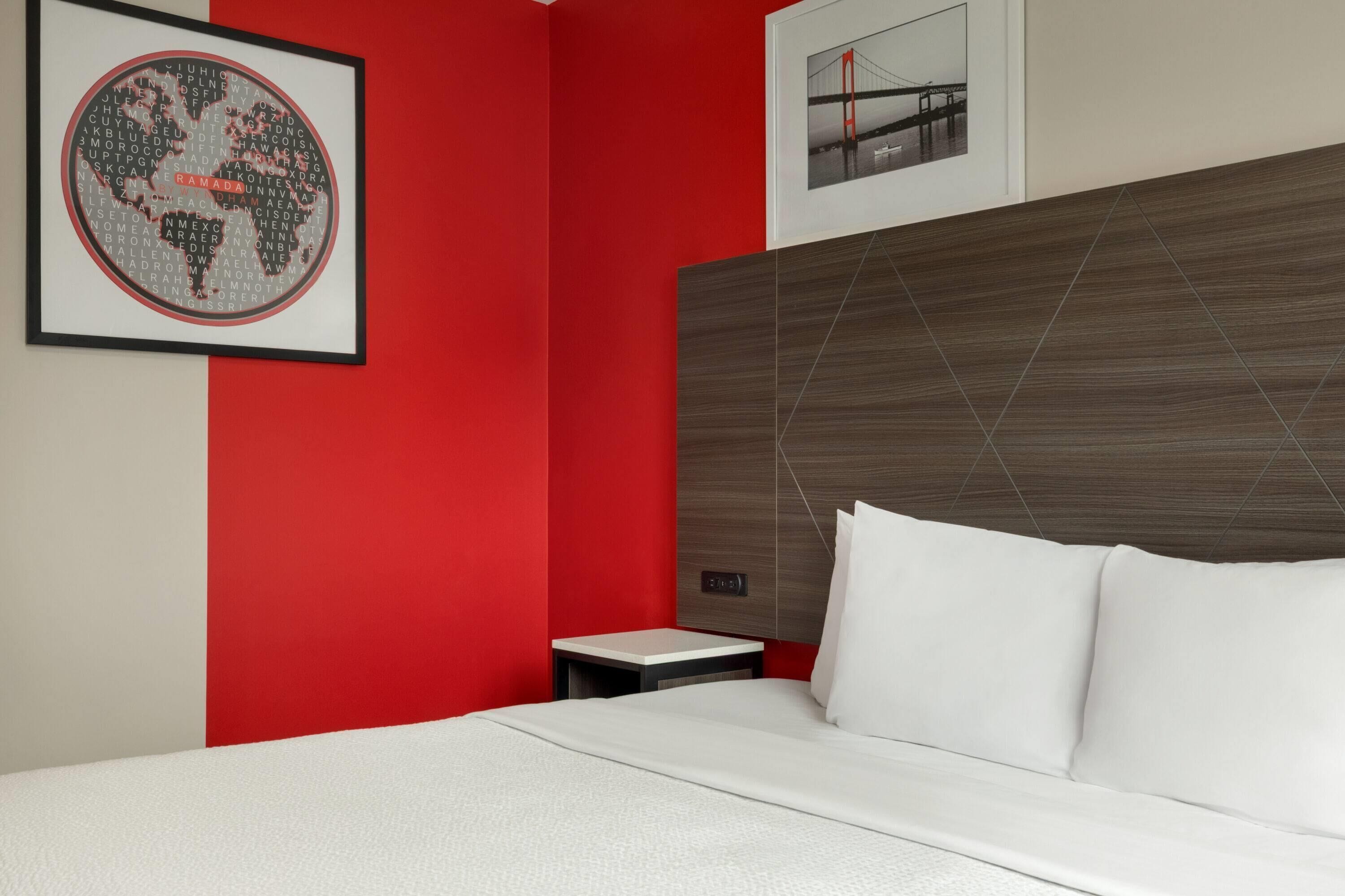 A hotel room with a red accent wall, a framed world map, a bed with white bedding, a small bedside table, and a framed picture of a bridge.