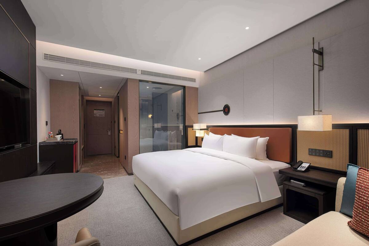 Ein modernes Hotelzimmer mit einem großen Bett, einem Schreibtisch, einem Stuhl, einem Fernseher und einem an der Wand montierten Telefon.