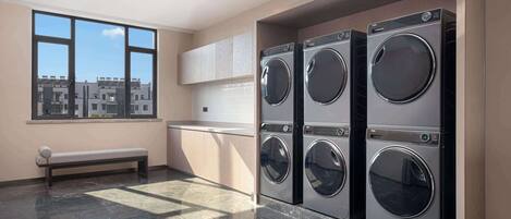 A laundry room with four front-loading washing machines, a bench, and a large window offering a view of buildings.