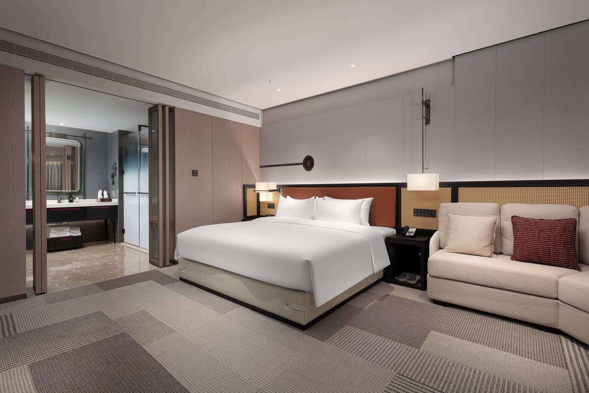 Ein modernes Hotelzimmer mit einem großen Bett, einer Couch, einem Schreibtisch und einem durch eine offene Tür einsehbaren Badezimmer.