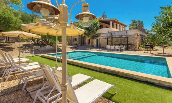 A swimming pool with lounge chairs, a pergola with umbrellas, and a stone building in the background.