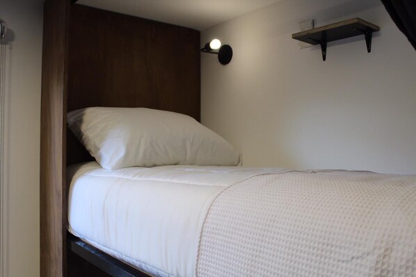 A single bed with a white mattress and pillows, a wooden headboard, a wall-mounted shelf with a light, and a plain white wall.
