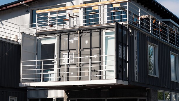 A multi-story building with a balcony on the upper level, large windows, and a visible shipping container on the ground floor.