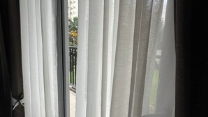 A view through a window with sheer curtains, revealing a balcony with a railing and a building in the background.