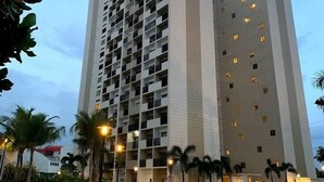 A tall, modern building with multiple floors and balconies, surrounded by greenery and palm trees.