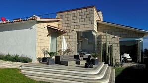 Une maison moderne avec une façade en pierre, un patio couvert agrémenté de mobilier d’extérieur et une vue dégagée sur l’intérieur grâce à de grandes portes vitrées.