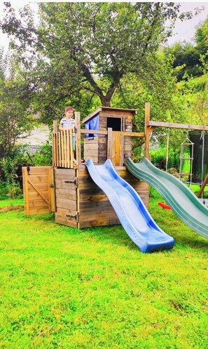 Explore a whimsical garden play area featuring a charming wooden house, slide, and swing set amidst lush greenery.