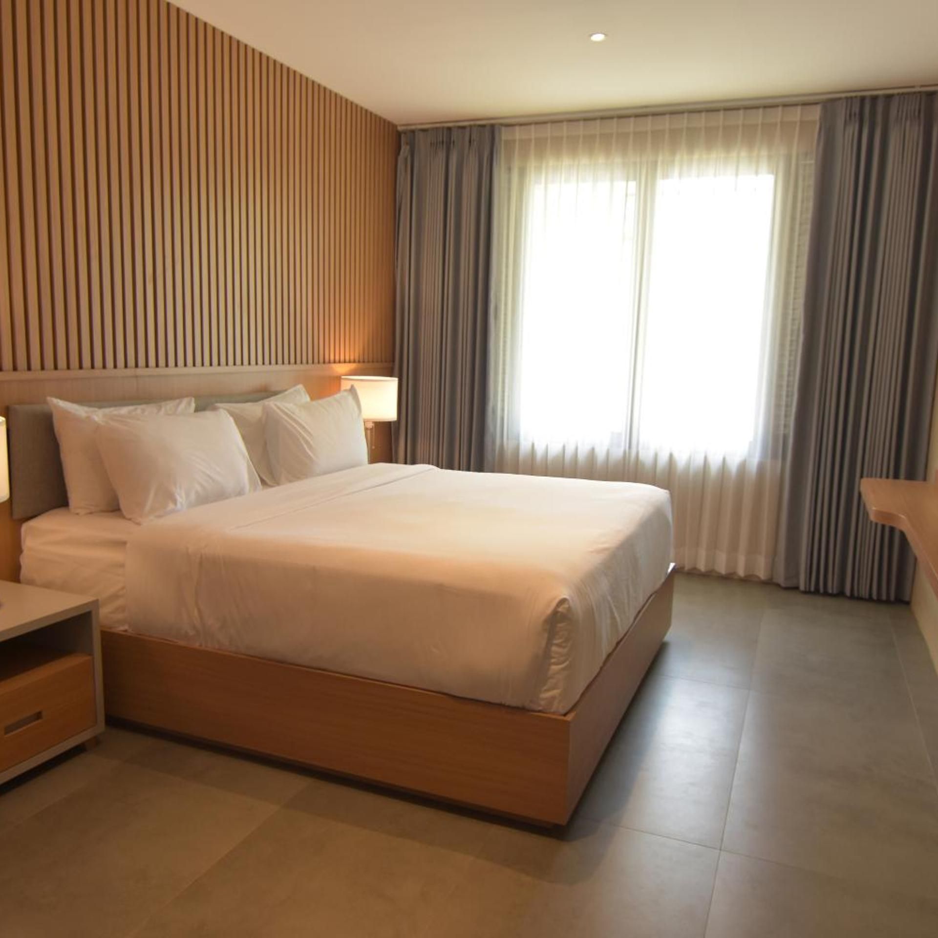Una habitación de hotel moderna con una cama grande, una mesita de noche y un ventanal con cortinas.