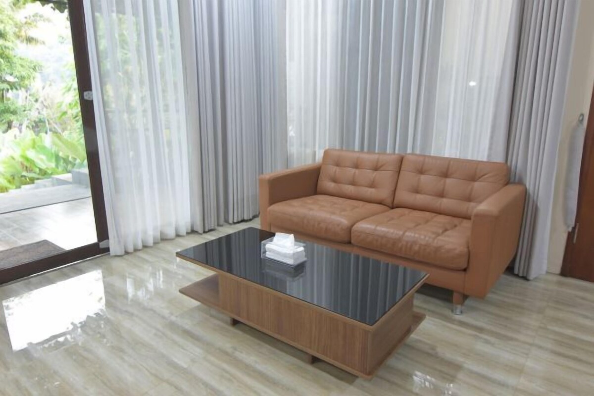 A modern living room with a leather sofa, a glass coffee table, and a view through sheer curtains.