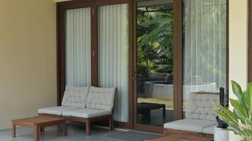 A balcony with two cushioned chairs and a wooden table, large sliding glass doors, and a view of a garden.