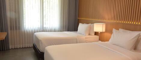 Habitación de hotel con dos camas, un ventanal grande con cortinas, un cabecero de madera y una lámpara de noche.