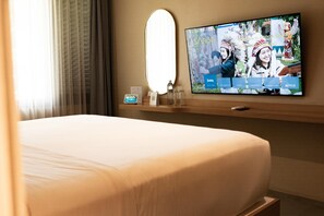 A hotel room with a bed, a TV mounted on the wall, a bedside table with a bottle and glasses, and a window with curtains.