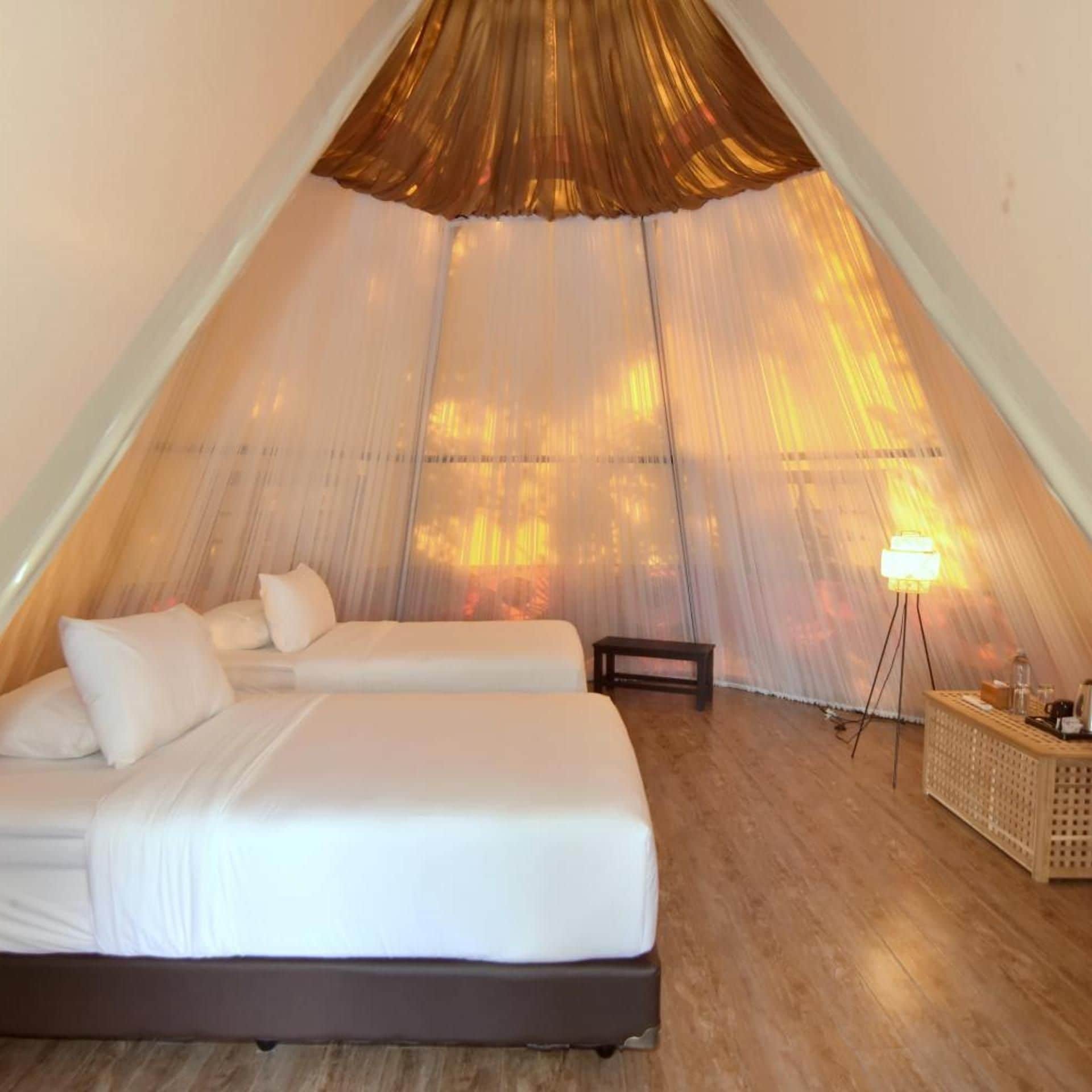 Quarto tipo tenda com duas camas, uma mesinha de cabeceira e um abajur.
