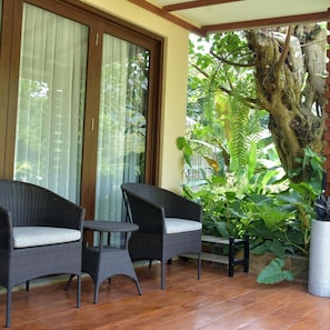 A covered patio with wicker furniture, a large tree, and a view of a lush garden. - Sarga Earthing Resort (Cipanas)