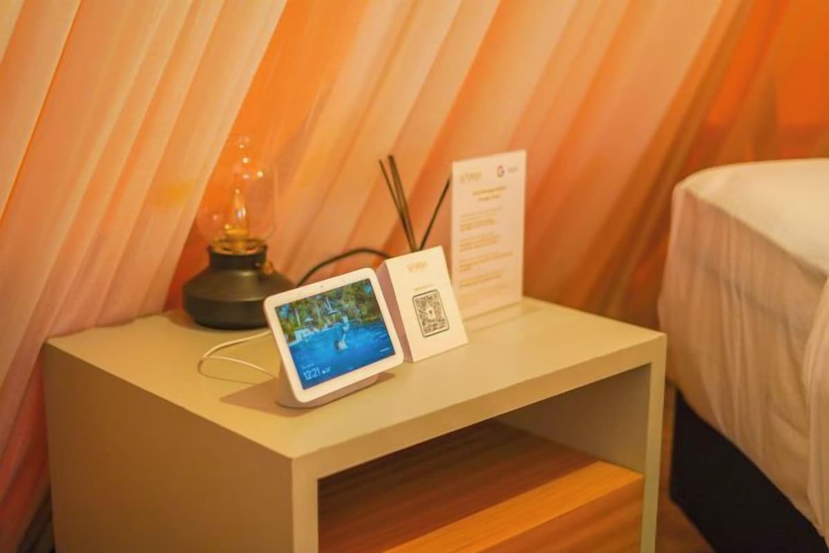 Habitación de hotel con una mesita de noche que incluye una tableta mostrando una imagen panorámica, una lámpara pequeña y un cartel con información de la habitación.