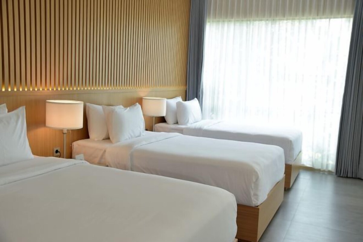 A hotel room with two beds, a wooden headboard, a nightstand with a lamp, and a window with curtains.