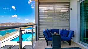 En balkon med udsigt over swimmingpool og havet, en blå sofa og et lille bord med et glas drik.