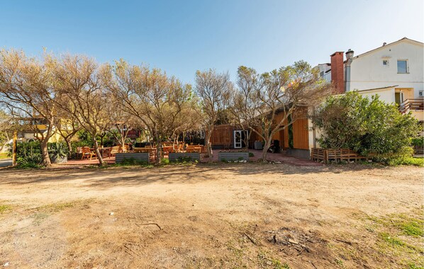 A rustic outdoor seating area with wooden furniture and a small building, surrounded by trees and a dirt path.